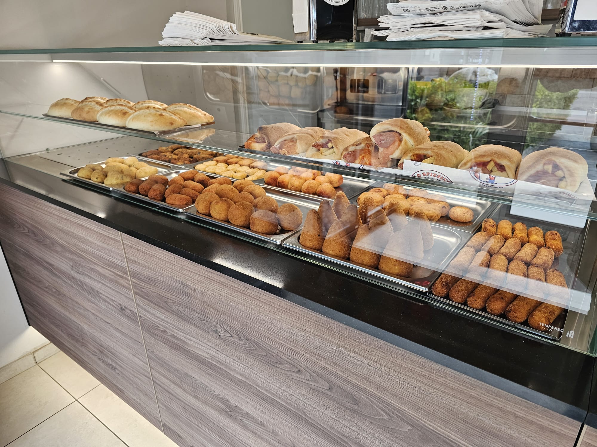 Vetrina di Lo Spizzico con arancini, crocchette, panini e calzoni farciti su vassoi d'acciaio, bancone in legno grigio