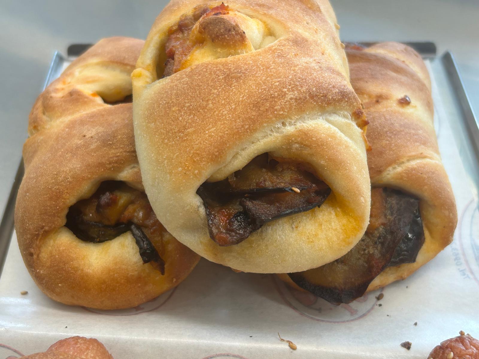 Tre rustici di pasta lievitata dorata ripieni di melanzane fritte e sugo, su carta da forno in un vassoio d'acciaio