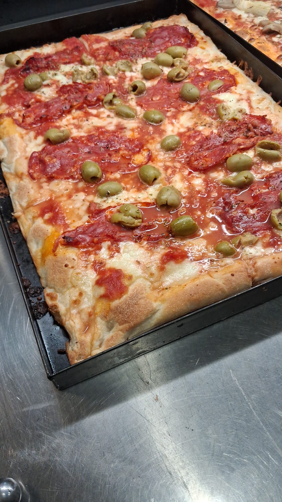 Teglia di pizza al taglio di Lo Spizzico con salame piccante, olive verdi e mozzarella su un banco d'acciaio
